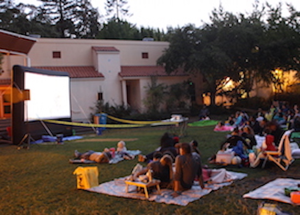 People gather on blankets in large backyard and watch movie on projection screen.